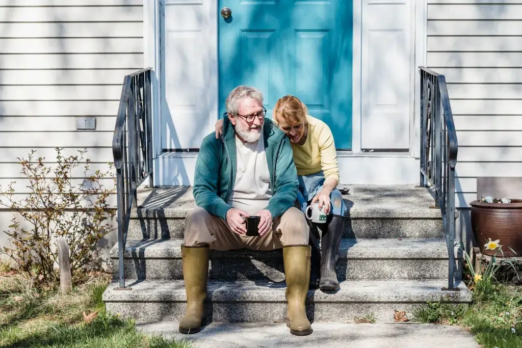An older couple sitting close together on their front steps in rain boots, holding mugs and talking